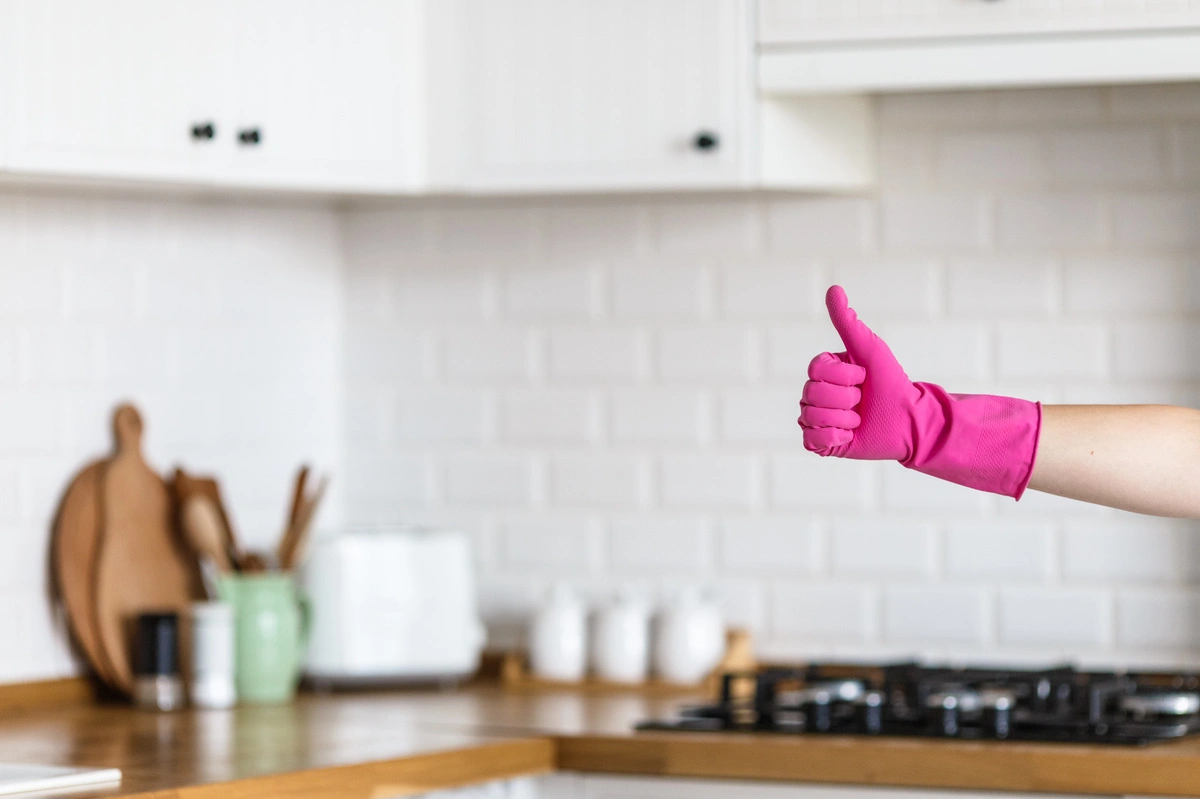 Nettoyage professionnel : Une main gantée de rose fait un signe de pouce en l'air devant une cuisine propre et bien rangée, symbolisant la satisfaction et la qualité du service.