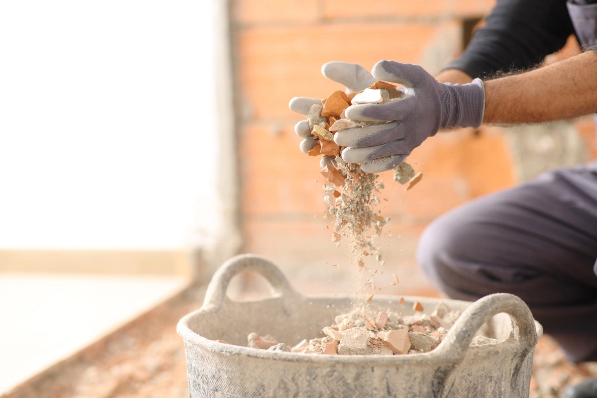 Nettoyage après sinistre : Un ouvrier en gants verse des débris de construction dans un seau, illustrant le processus de nettoyage après sinistre pour éliminer les restes de matériaux endommagés.